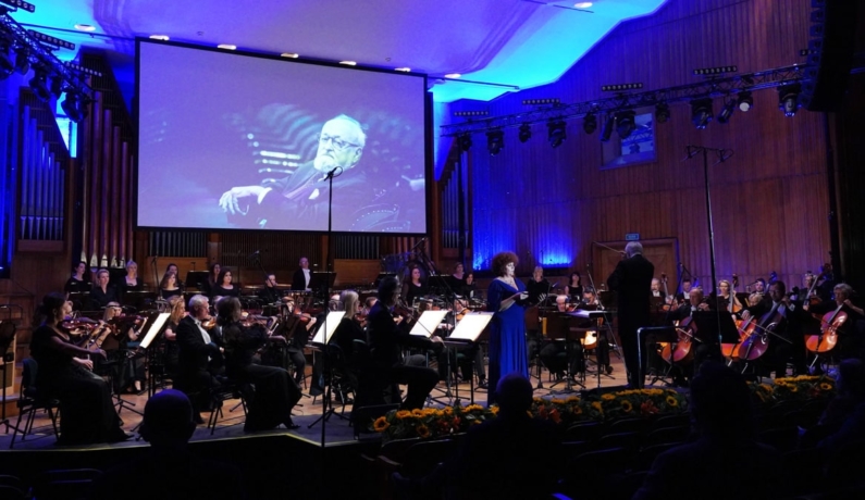 Orkiestra wraz z wokalistką stoją na scenie, w tle ekran wyświetla portret Pendereckiego