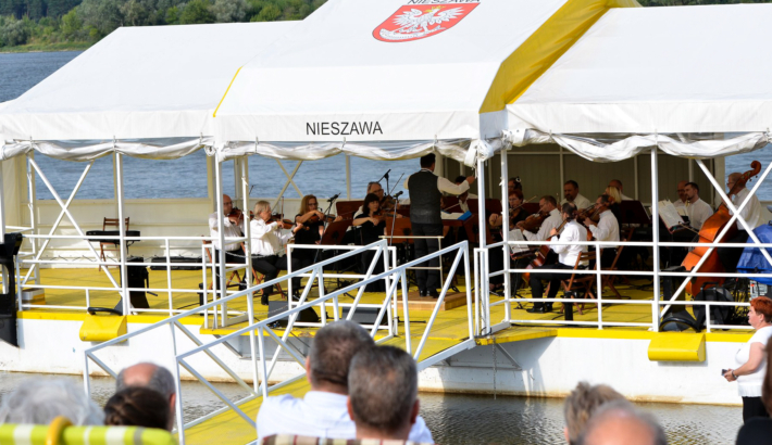 Na pokładzie przycumowanego promu muzycy grają na instrumentach, na brzegu melomani słuchają muzyki