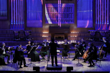 zdjęcie orkiestry kameralnej na przyciemnionej estradzie z wyświetlonym na prospekcie krzyżem