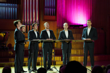 szóstka mężczyzn w czarnych garniturach stoi przy pulpitach i śpiewa