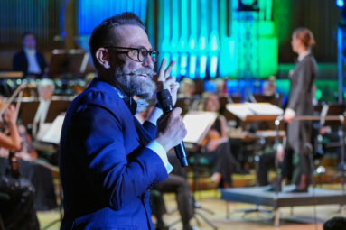 zdjęcie mężczyzny w okularach, z siwym wąsem, przemawiającego do publiczności przy z mikrofonem w ręku