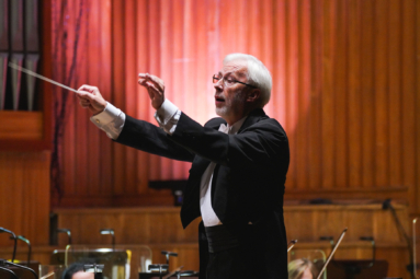 zdjęcie starszego siwego mężczyzny z uniesionymi rękami zwróconymi w stronę orkiestry