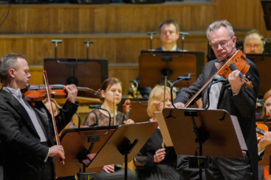 zdjęcie dwóch mężczyzn we frakach i białych muszkach. Jeden z nich gra na altówce, a drugi na skrzypcach, w tle siedzą muzycy orkiestry symfonicznej