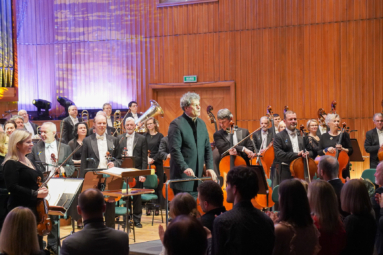 zdjęcie dyrygenta i muzyków orkiestry stojących na estradzie, uśmiechających się do oklaskującej ich publiczności