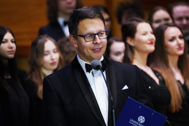 zdjęcie mężczyzny w średnim wieku, w granatowych okularach przemawiającego do mikrofonu