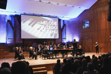 zdjęcie estrady na której występują artyści muzycy a za nimi wyświetlony jest duży ekran z tytułem koncertu i obrazkiem przedstawiającym kalwisze fortepianu