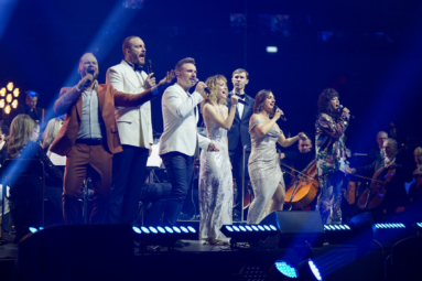 zdjęcie sześciu wokalistów ubranych w eleganckie stroje śpiewających do mikrofonów z towarzyszeniem orkiestry