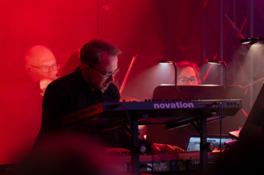 Muzyk grający na instrumentach klawiszowych podczas koncertu, otoczony czerwonym oświetleniem scenicznym. W tle widać orkiestrę.