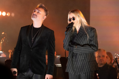 Dwóch wokalistów na scenie – mężczyzna w czarnym garniturze i kobieta w ciemnym, prążkowanym stroju oraz okularach przeciwsłonecznych. W tle członkowie orkiestry.