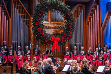 Chór i orkiestra podczas świątecznego koncertu. Chór chłopięcy i męski, ubrany w czerwone i czarne stroje, śpiewa na tle organów ozdobionych wielkim świątecznym wieńcem. Przed nimi gra orkiestra.