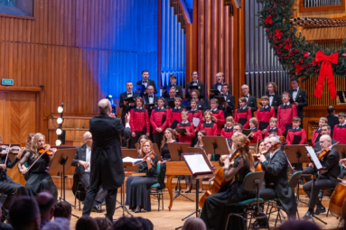 Dyrygent w czarnym fraku energicznie prowadzi koncert, orkiestra i chór śpiewają pod jego batutą.