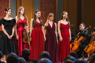 cztery młode dziewczyny w długich sukienkach koncertowych uśmiechające się w strone publiczności