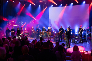 Estrada z zespołem, orkiestrą i efektownym oświetleniem. Publiczność ogląda występ na żywo.