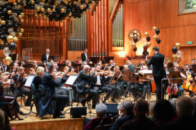 Orkiestra symfoniczna gra w sali koncertowej ozdobionej złoto-czarnymi balonami. Dyrygent prowadzi muzyków, a publiczność obserwuje koncert.