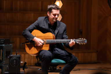 mężczyzna w czarnym garniturze i czarnej koszuli grający na gitarze podłączonej do wzmacniacza stojącego obok