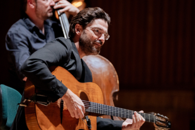 mężczyzna w gęstych, ciemnych włosach i okularach grający na gitarze i spoglądający z uśmiechem w stronę publiczności