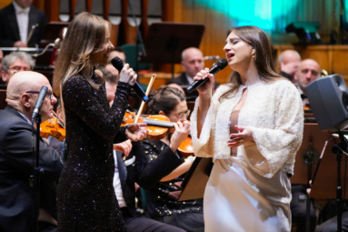 Dwie śpiewaczki w eleganckich kreacjach wykonują utwór na tle orkiestry symfonicznej. Widać emocje na ich twarzach