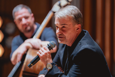 mężczyzna w siwych włosach przemawiający przez mikrofon. Na lewej ręce ma założony zegarek, za nim w tle przysłuchuje się kontrabasista