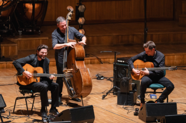 Trzech mężczyzn ubranych na czarno, dwóch z nich siedzi na krzesłach i grają na gitarze, a trzeci stpo i gra na kontrabasie. Wszyscy się uśmiechają