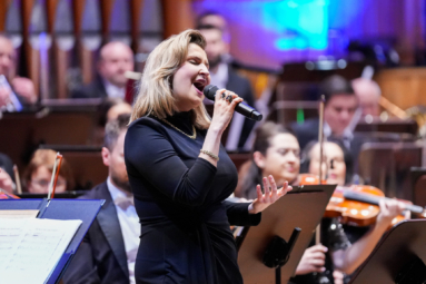 Wokalistka w czarnej sukni śpiewa na tle orkiestry symfonicznej.