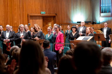 młode kobiety uśmiechające się do oklaskującej je publiczności, obok nich stoi orkiestra i uśmiecha się w ich stronę