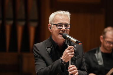 siwy mężczyzna w okularach z czarnymi oprawkami, śpiewający do mikrofonu ustawionego na statywie