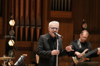 siwy mężczyzna w okularach z czarnymi oprawkami, śpiewający do mikrofonu ustawionego na statywie